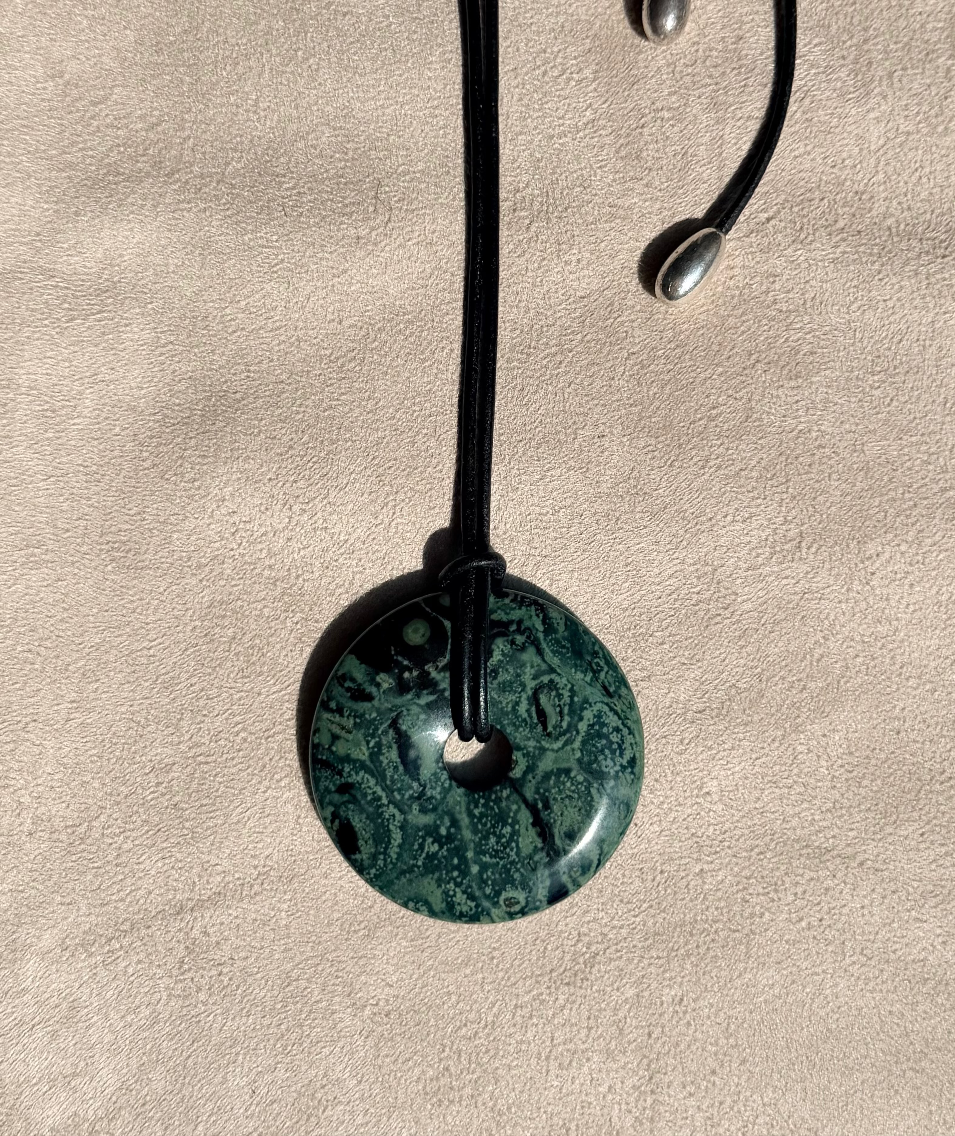 A round green kambaba stone pendant necklace with a black cord and silver-finish displayed on a textured beige surface.