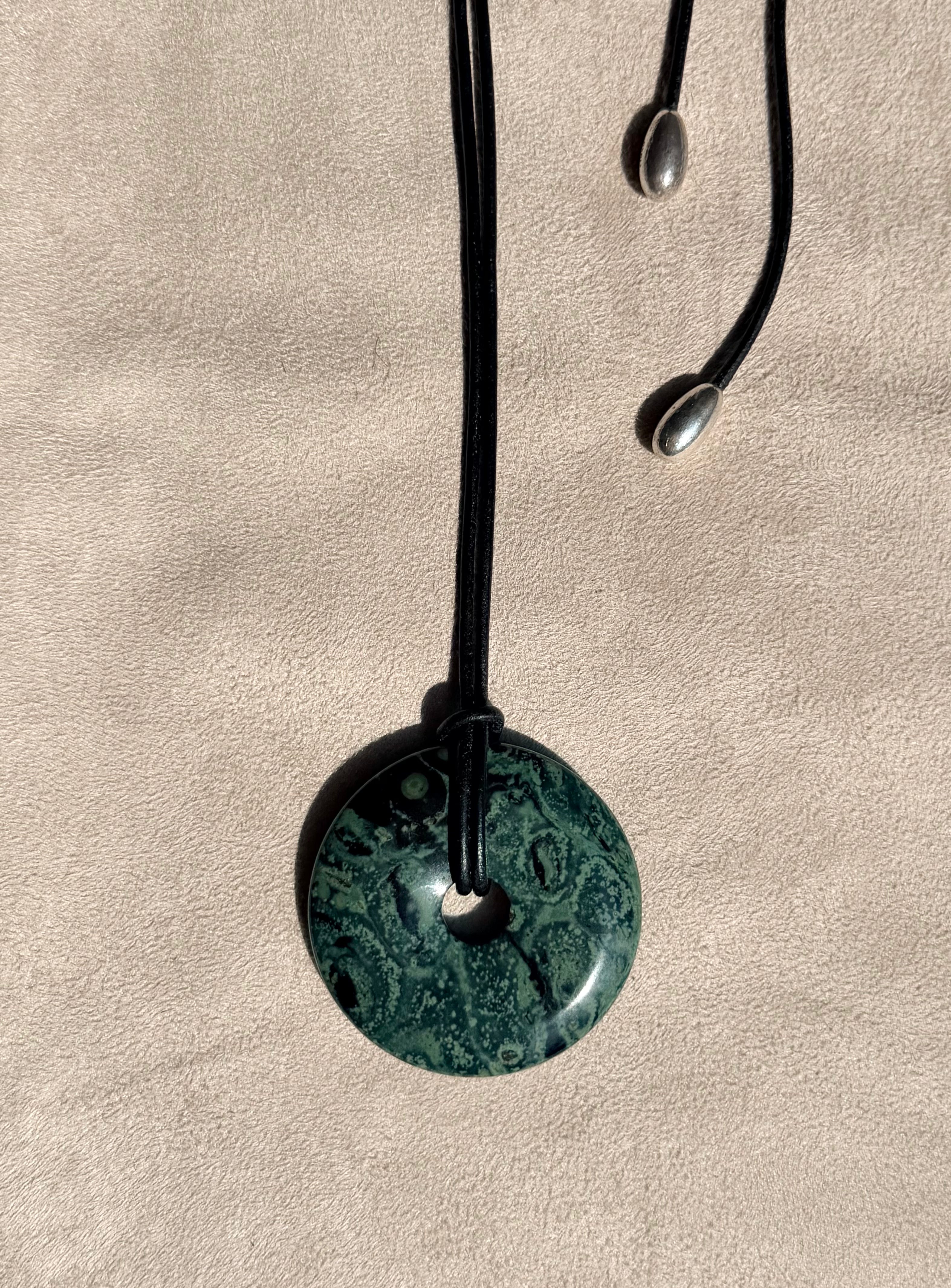 A round green kambaba stone pendant necklace with a black cord and silver-finish displayed on a textured beige surface.
