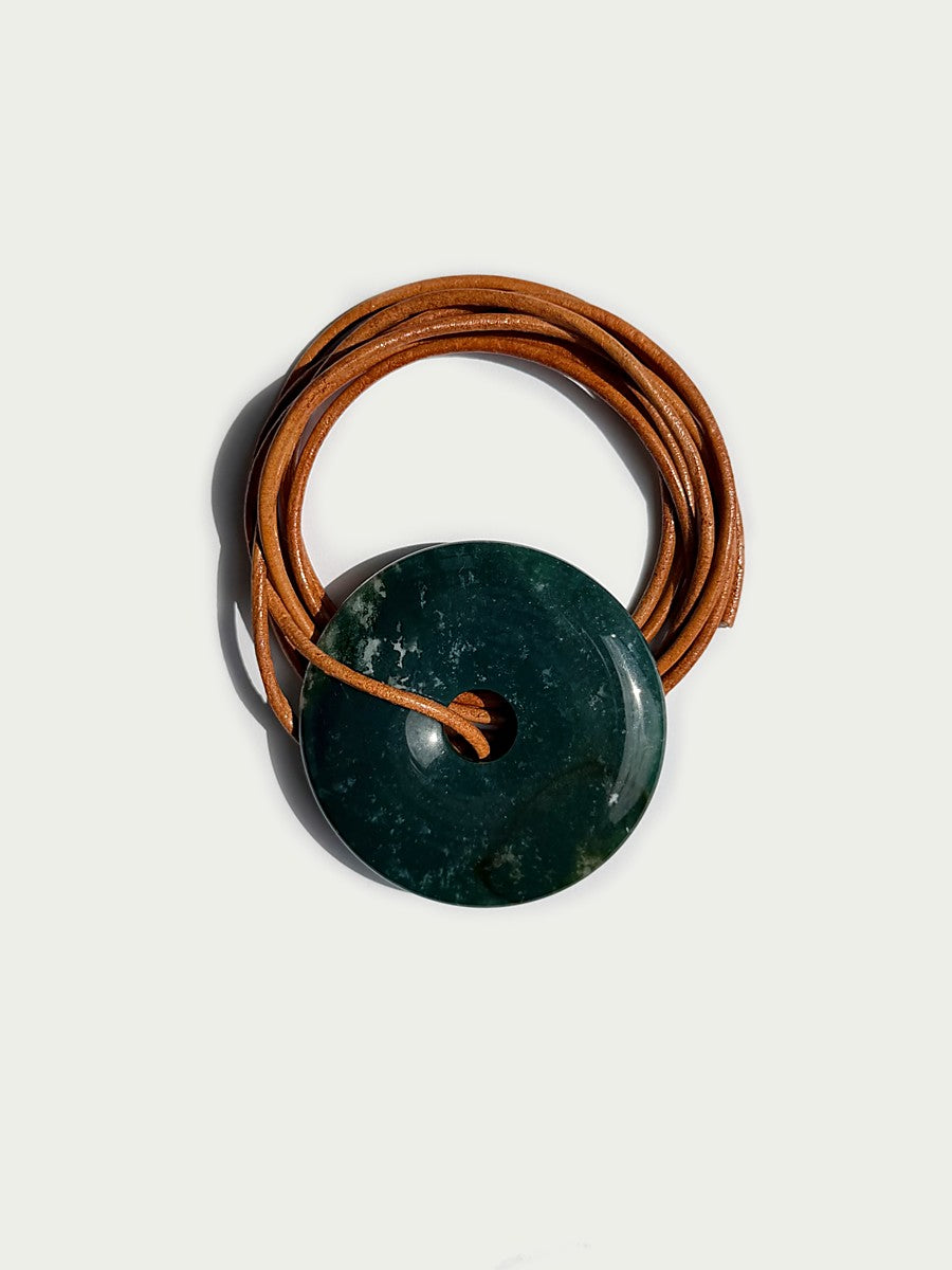 A dark green polished stone pendant with a natural marbled texture, threaded onto a coiled brown leather cord, photographed on a light off-white background.