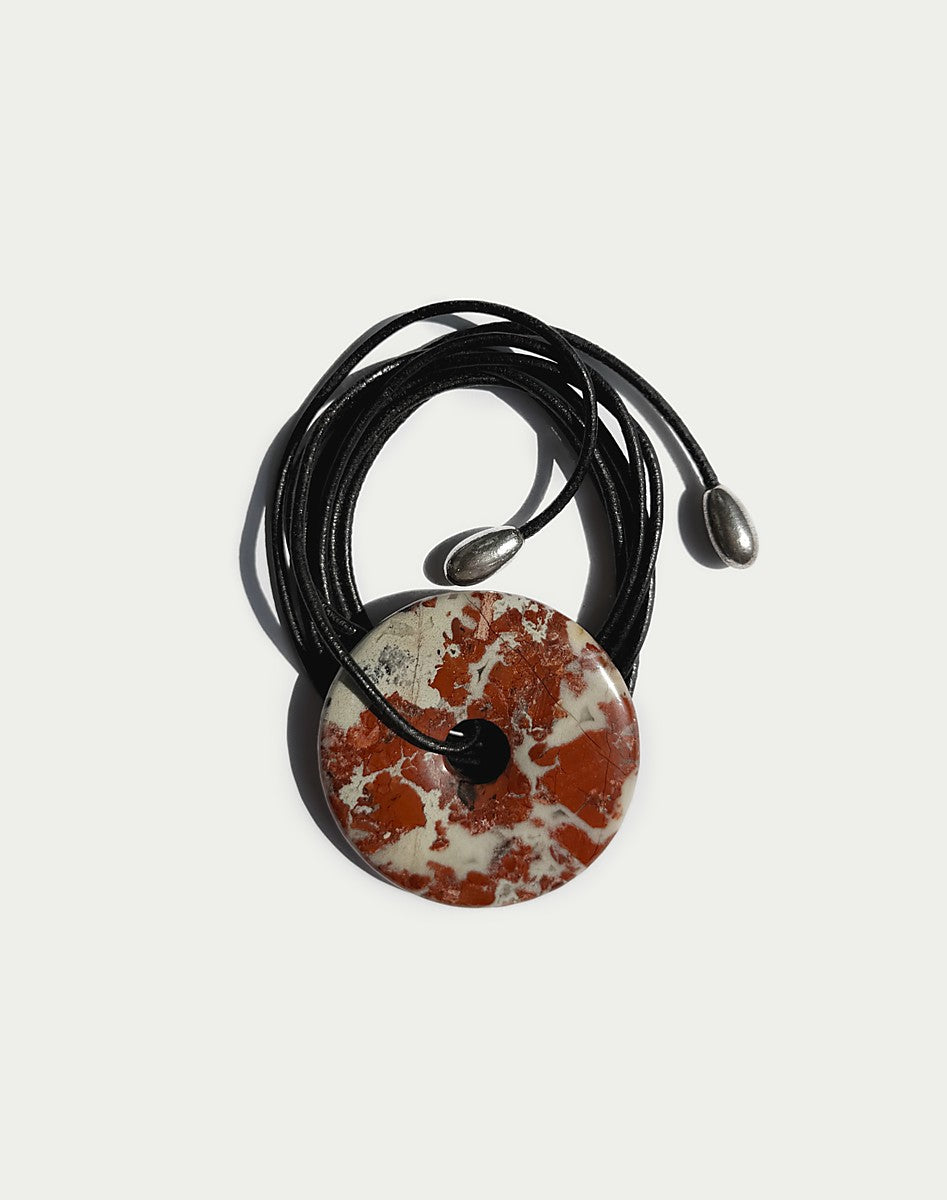 A round pendant necklace featuring a red and white marbled stone disc on a black cord, with small silver metal beads at the ends, shown against a light gray background.
