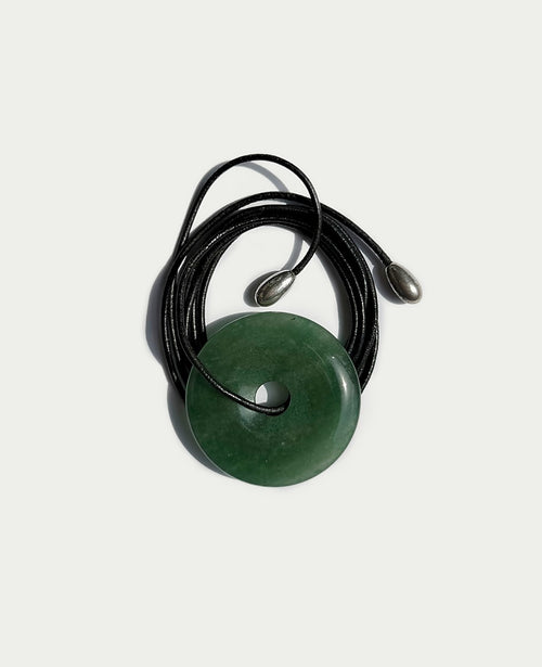 A green circular stone pendant with a central hole, threaded on a black cord necklace with silver-toned beads, set against a light gray background.