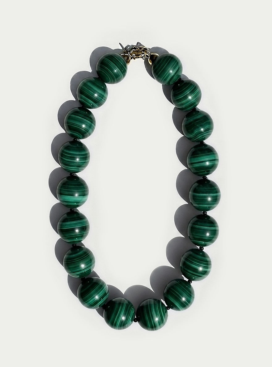 A beaded necklace made of polished green malachite beads with natural dark green stripes, arranged in an oval shape on a light gray background.